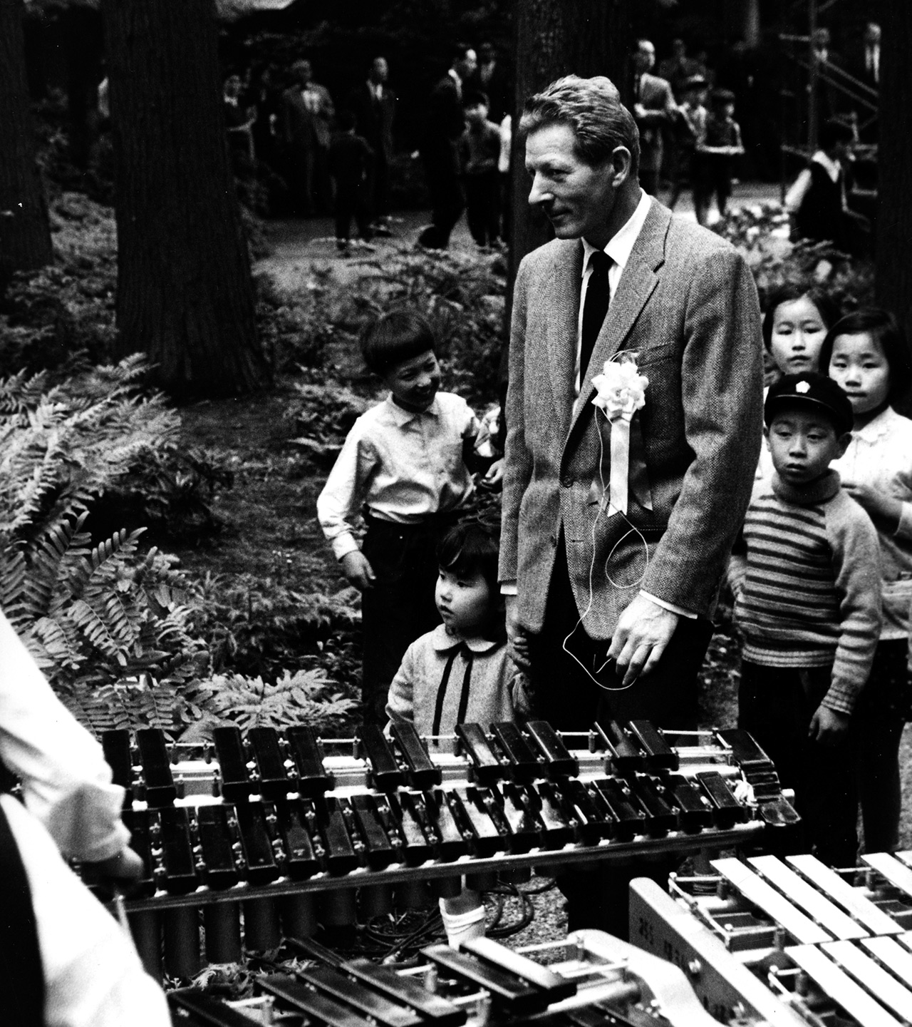 Image 1 of [ Danny Kaye, while on tour of Japan and six Asian nations, stands surrounded by children amidst trees in Japan? near xylophones?]
